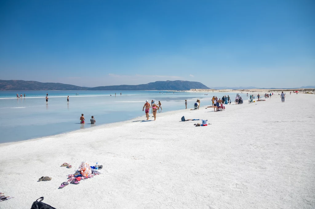 Salda to jedno z najczystszych jezior w Turcji, jednak od kilku lat nie można się w nim kąpać Szeroka, biała plaża nad turkusowym jeziorem Salda, otoczona przez kąpiących się ludzi i grupki spacerowiczów, w tle rozciąga się górzysty krajobraz.