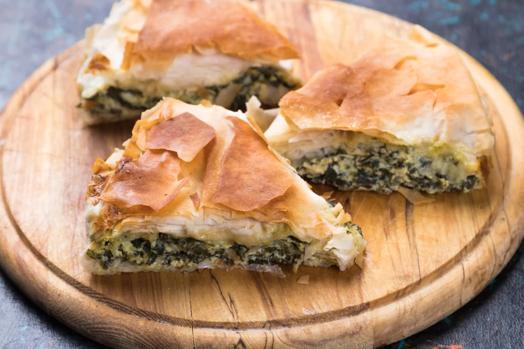 Burek ze szpinakiem i fetą, po grecku - spanakopita Trzy kawałki placka z ciasta filo z nadzieniem ze szpinaku i białego sera leżą na okrągłej, drewnianej desce.