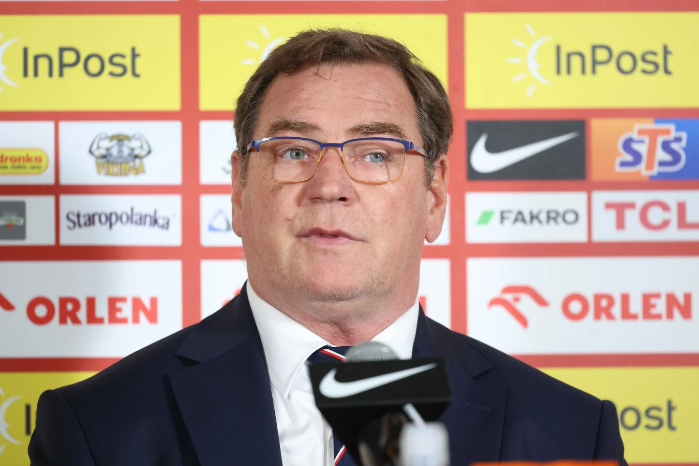 Jan Urban Mężczyzna w garniturze i okularach wypowiada się na tle ściany z logotypami sponsorów podczas konferencji prasowej.