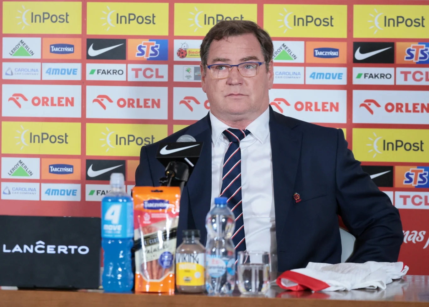 Mężczyzna w garniturze siedzi przy stole podczas konferencji prasowej, za nim widoczna jest ścianka z logo sponsorów, na stole stoją butelki z napojami, mikrofon i materiały reklamowe.