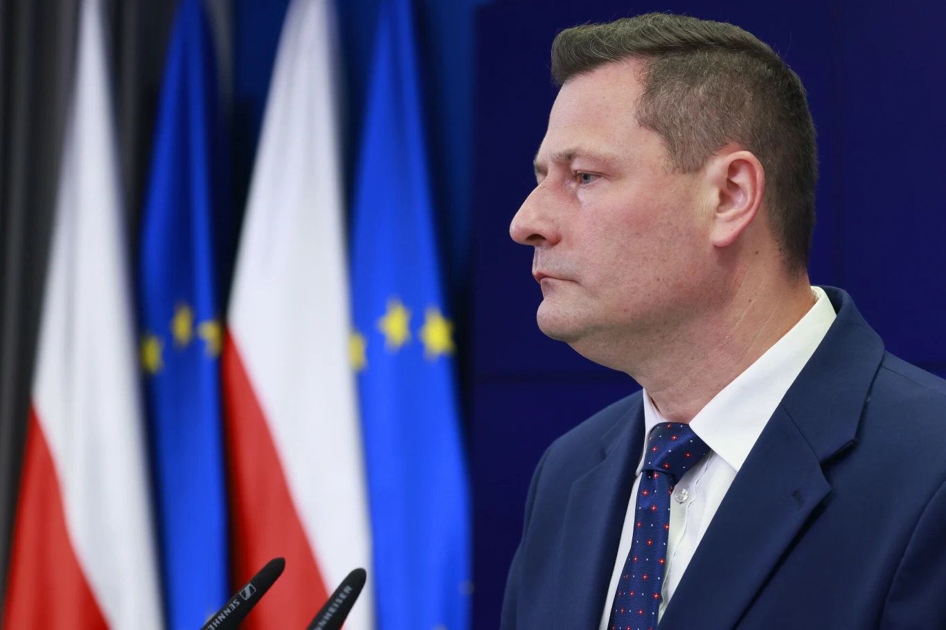 Mężczyzna w garniturze i krawacie stoi przy mównicy, w tle widoczne flagi Polski oraz Unii Europejskiej.
