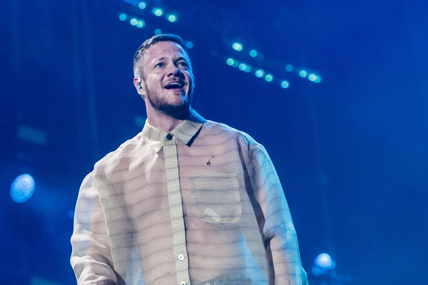 Imagine Dragons zagrają dwa koncerty w Warszawie Mężczyzna na scenie koncertowej ubrany w przezroczystą koszulę, oświetlony niebieskim światłem, w tle rozmyte reflektory i elementy sceniczne.