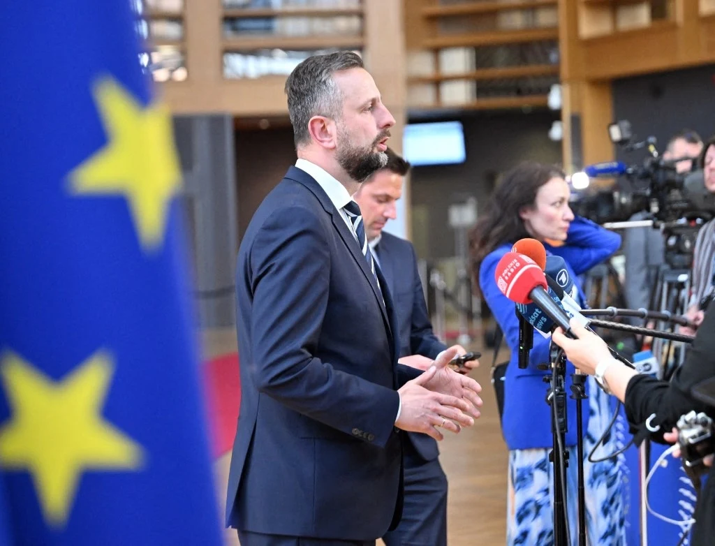 Mężczyzna w garniturze udziela wywiadu dziennikarzom, w tle widoczne są kamery, mikrofony oraz fragment flagi Unii Europejskiej.