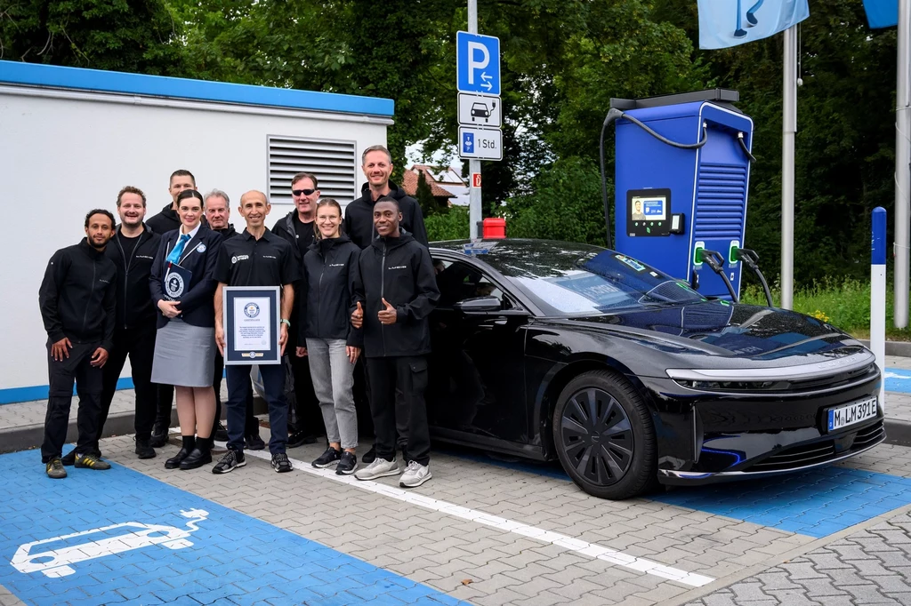 Lucid Air Grand Touring pobił rekord Guinnessa. Grupa osób uśmiecha się i pozuje obok czarnego samochodu elektrycznego stojącego przy stacji ładowania, jedna z nich trzyma certyfikat, w tle widoczne niebieskie oznaczenia dla pojazdów elektrycznych.