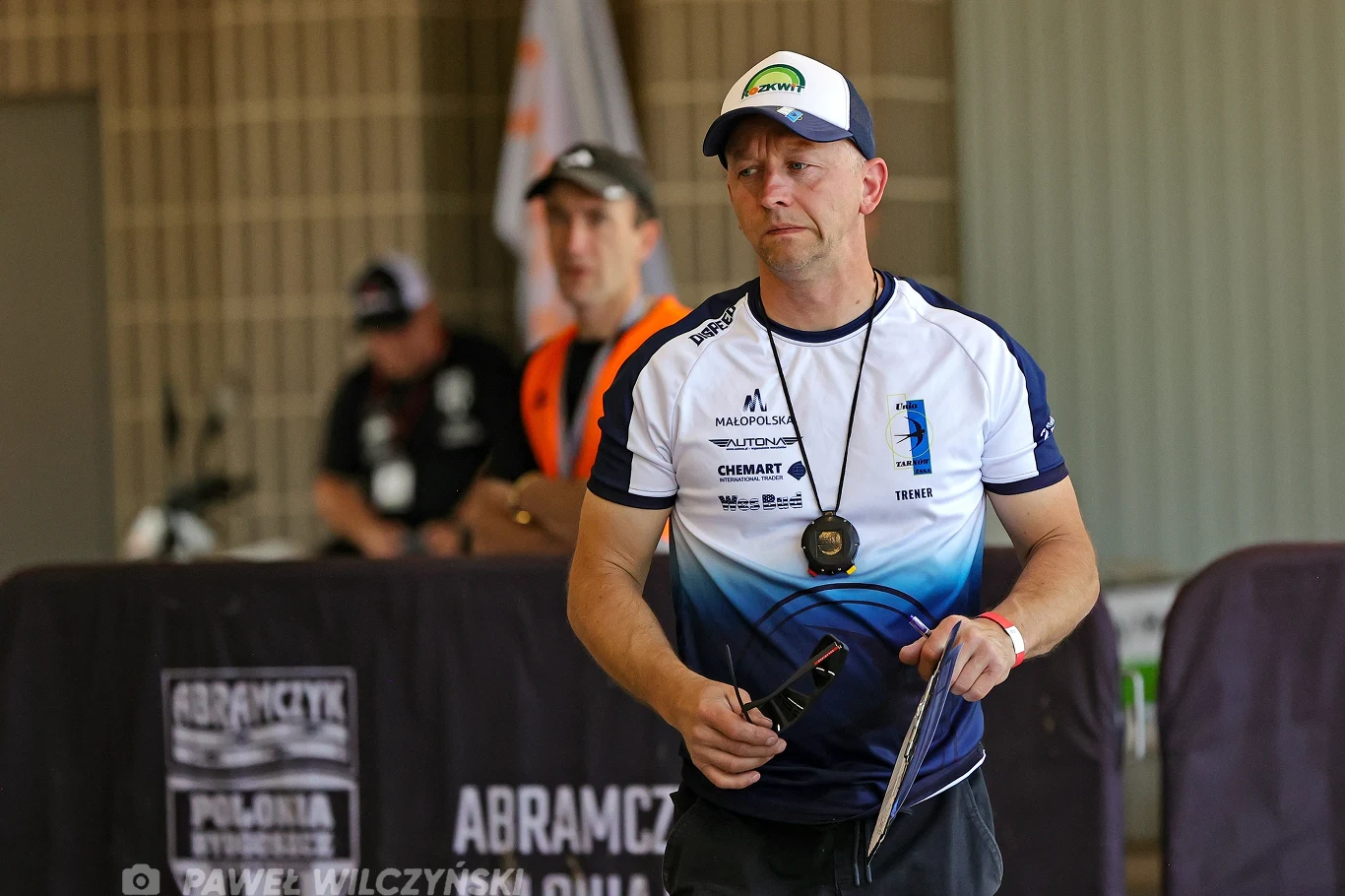 Mężczyzna w sportowej koszulce i czapce z daszkiem trzyma gwizdek oraz kartkę, ma zawieszony medal na szyi, w tle widoczna druga osoba oraz banery z nazwami organizacji