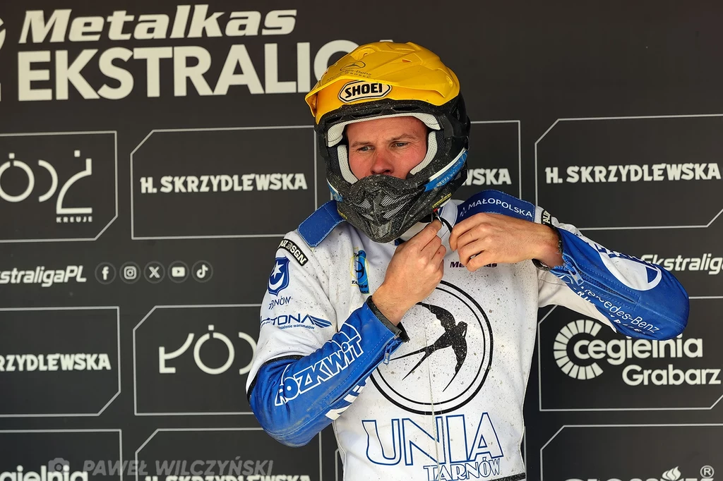 Matej Zgar Żużlowiec w stroju ochronnym zakłada kask, na tle reklam sponsorów i tablicy z nazwą ligi żużlowej, ubrany w biało-niebieski kombinezon z emblematem UNIA Tarnów.