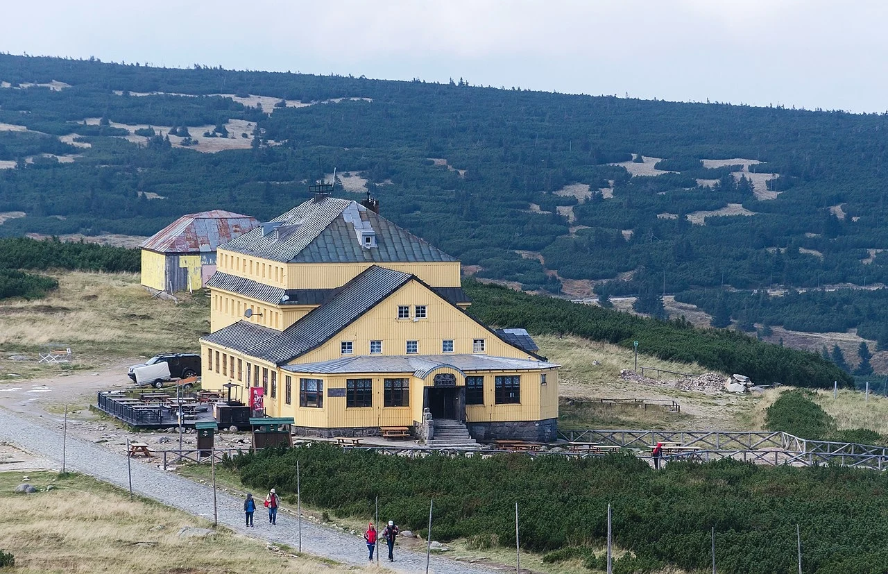 Duży, żółty budynek schroniska górskiego położony w otoczeniu niskiej roślinności i górskich stoków, z widoczną drogą prowadzącą w jego kierunku oraz kilkoma spacerującymi osobami.