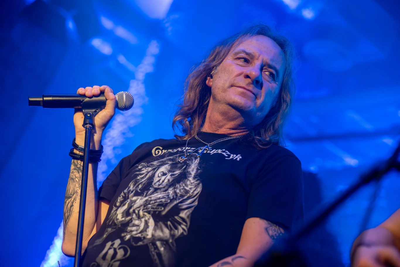 Grzegorz Kupczyk uznawany jest za legendę polskiego heavy metalu Mężczyzna z długimi włosami trzyma mikrofon, ubrany w czarną koszulkę z nadrukiem grafiki, stoi na scenie koncertowej wśród niebieskiego oświetlenia.