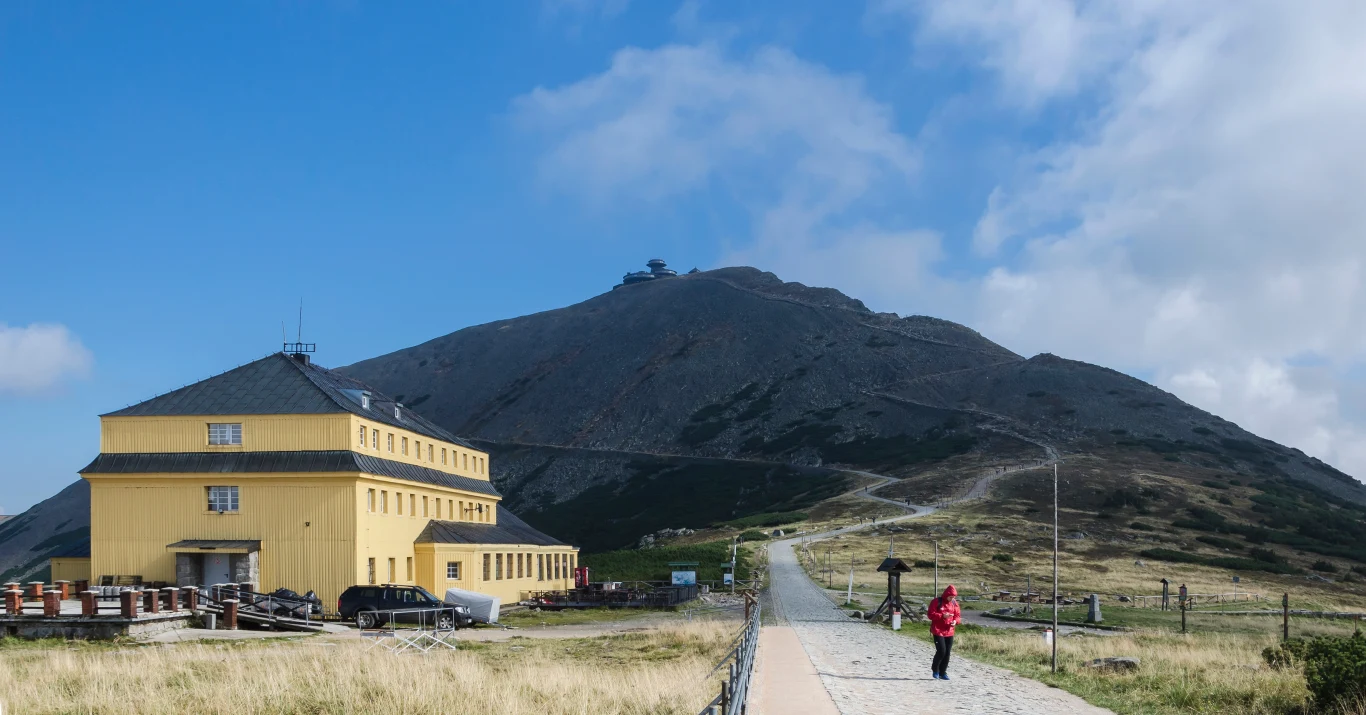 Żółty budynek schroniska stoi u podnóża góry, obok biegnie brukowana ścieżka, po której spaceruje osoba w czerwonej kurtce, w tle góra z charakterystycznym budynkiem na szczycie pod błękitnym niebem z lekkimi chmurami.