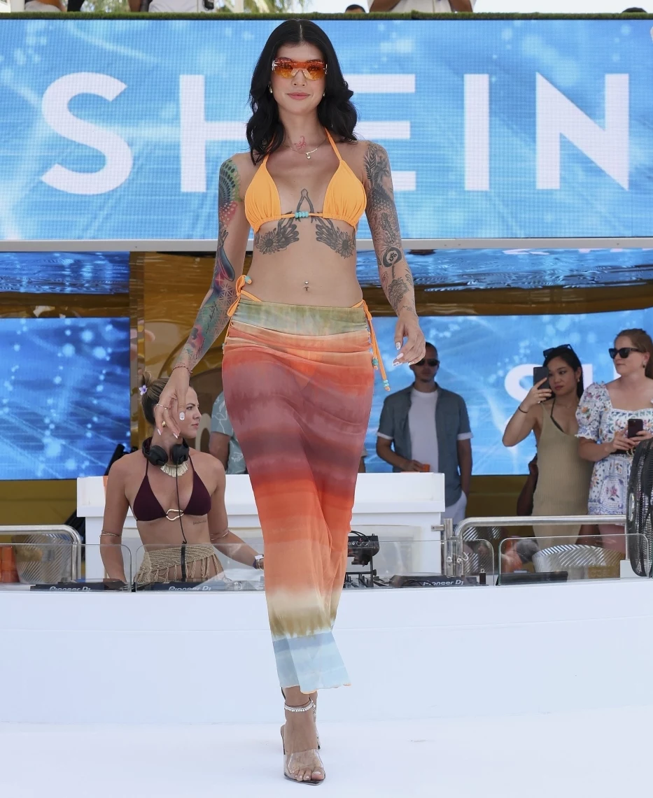 SHEIN otwiera sezon letni hucznym pokazem mody na Ibizie Modelka o tatuażach prezentuje kolorowy strój plażowy, stojąc na wybiegu podczas pokazu mody na tle logotypu SHEIN, w otoczeniu widowni i organizatorów.