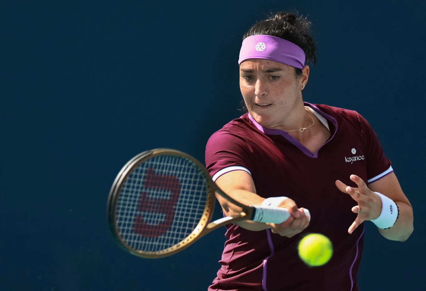 Tenisistka wykonuje zamach rakietą do tenisa, skupiona na odbiciu piłki, ubrana w bordowy strój sportowy i fioletową opaskę na głowie, jasne tło kortu.