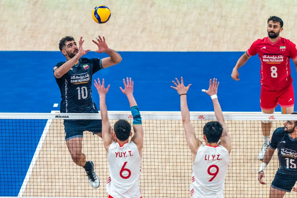 Liga Narodów siatkarzy. Kadr z meczu Chiny - Iran Gracz drużyny w czarnych strojach wykonuje atak nad siatką, podczas gdy dwóch zawodników drużyny przeciwnej w białych koszulkach z numerami 6 i 9 stara się zablokować piłkę podczas meczu siatkówki na hali sportowej