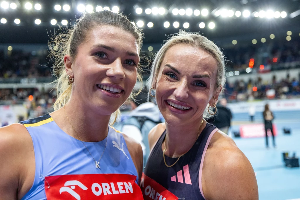 Kornelia Lesiewicz (po lewej) i Justyna Święty-Ersetic Dwie uśmiechnięte zawodniczki w sportowych strojach stoją obok siebie na tle jasnych świateł hali lekkoatletycznej, z logotypem Orlen na ich numerach startowych.