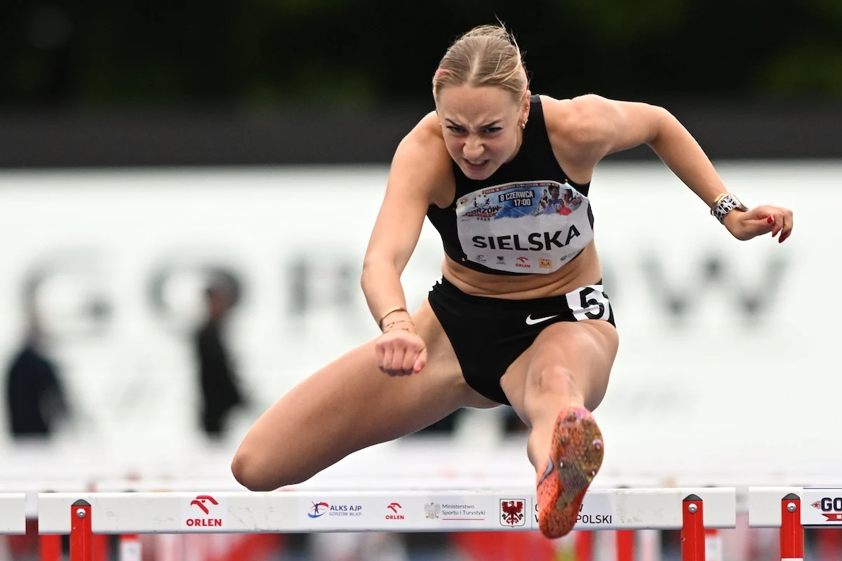 Alicja Sielska Lekkoatletka w trakcie biegu przez płotki, dynamiczna poza w trakcie pokonywania przeszkody, skupienie na twarzy, sportowy strój startowy z numerem startowym i nazwiskiem.