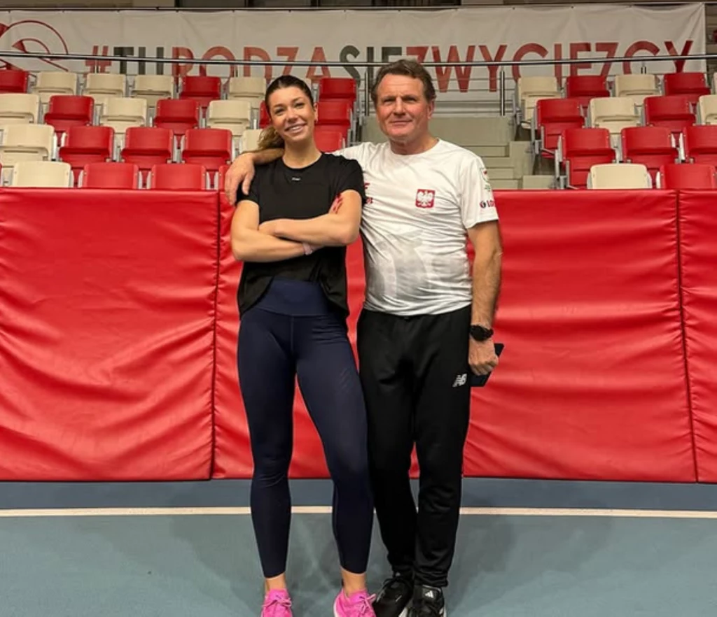 Kornelia Lesiesicz i trener Andrzej Giza Dwie osoby stojące przy sobie na tle czerwonej maty i pustych trybun, ubrani sportowo, uśmiechnięci, pozują do zdjęcia w hali sportowej.