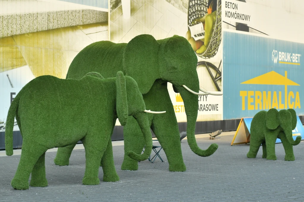 Trzy figury słoni pokryte zieloną sztuczną trawą ustawione na tle budynków i reklam, przestrzeń miejska w jasnym świetle dziennym.