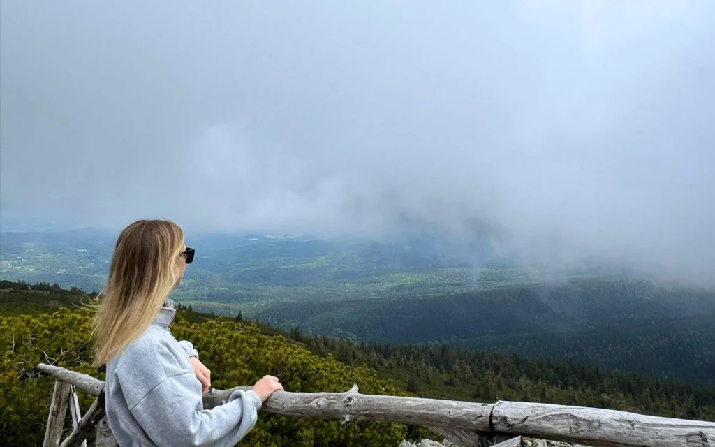 Karkonosze nawet zamglone robią ogromne wrażenie Kobieta w okularach przeciwsłonecznych patrzy z drewnianego ogrodzenia na rozległą, zieloną dolinę oraz góry przysłonięte mgłą; w tle widać szeroki krajobraz naturalny.