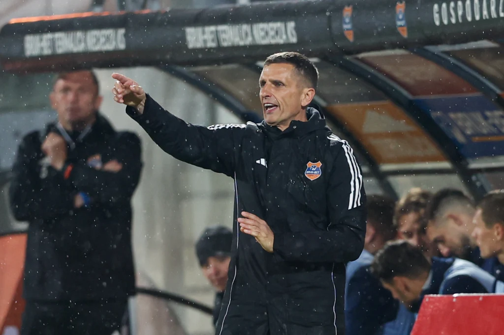 Trener drużyny piłkarskiej w czarnej kurtce wydaje polecenia stojąc przy linii bocznej boiska, za nim znajduje się ławka rezerwowych z zawodnikami i członkami sztabu.