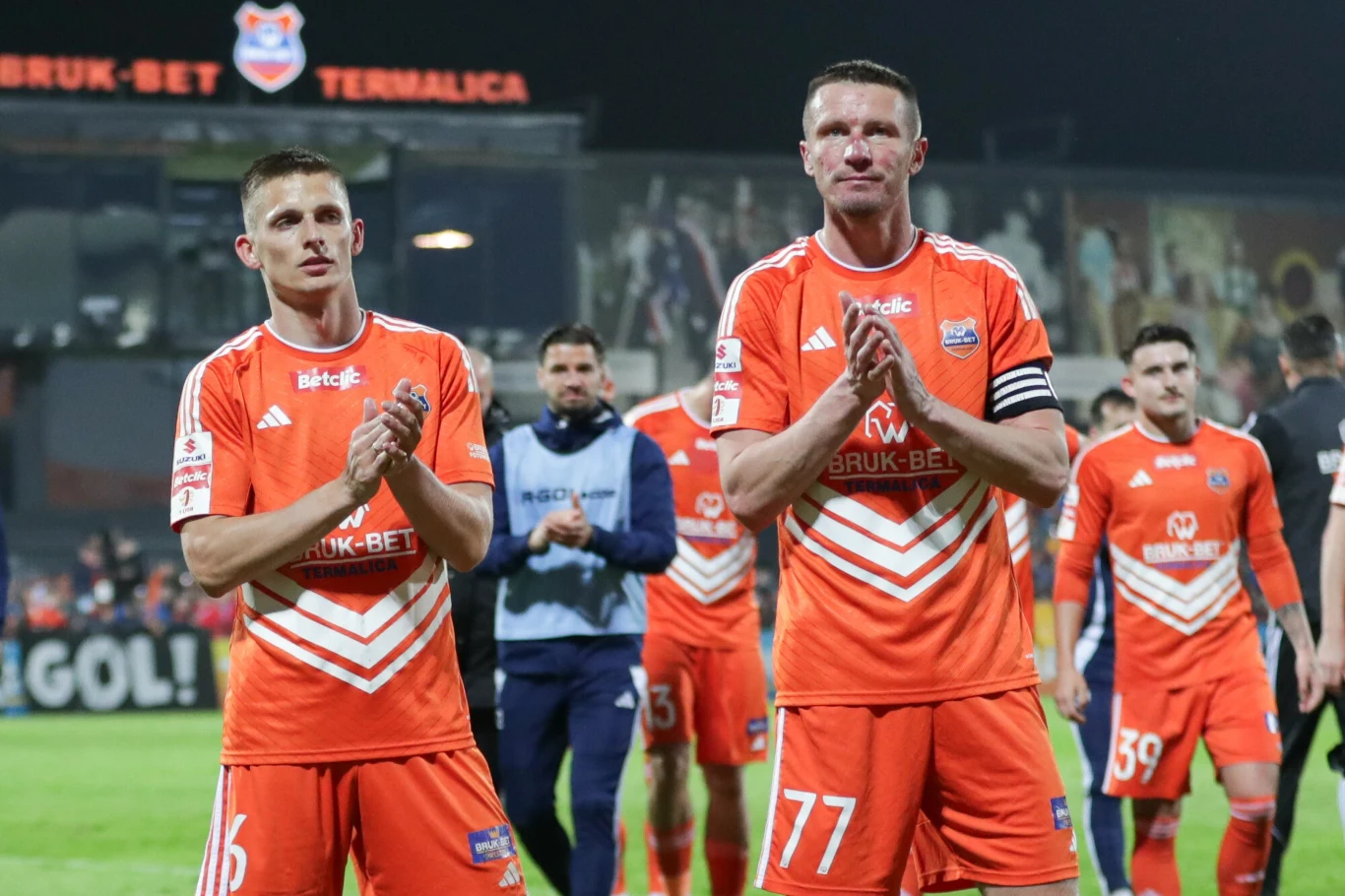 Piłkarze Bruk-Betu Termaliki Nieciecza miniony sezon Betclic I ligi zakończyli na drugim miejscu i mogli świętować powrót do Ekstraklasy Dwóch piłkarzy w pomarańczowych strojach drużyny Bruk-Bet Termalica bije brawo, za nimi widoczni są inni zawodnicy oraz kibice na trybunach stadionu, panuje wieczorna atmosfera sportowego wydarzenia.