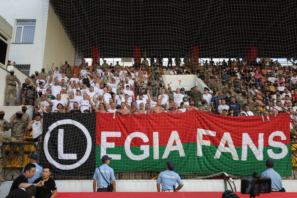 Fani warszawskiej Legii na Stadionie Centralnym w Aktobe Tłum kibiców Legii Warszawa ubrany w białe koszulki, stojący za dużą flagą z napisem 'Legia Fans', oddzielony od reszty stadionu siatką i pilnowany przez ochroniarzy oraz funkcjonariuszy.