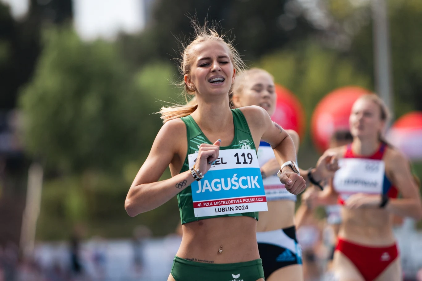 Biegaczka w zielonym stroju z widocznym numerem startowym kończy wyścig na bieżni, na tle innych zawodniczek, skupiona na wysiłku i rywalizacji podczas zawodów sportowych.