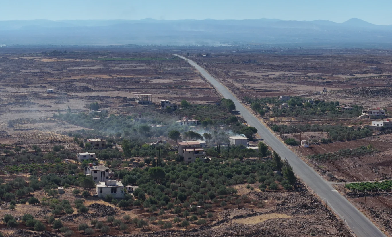 Prosta, asfaltowa droga przecinająca suchy krajobraz z niewielką liczbą domów otoczonych polami i roślinnością, delikatny dym unoszący się w kilku miejscach, wzgórza na horyzoncie.