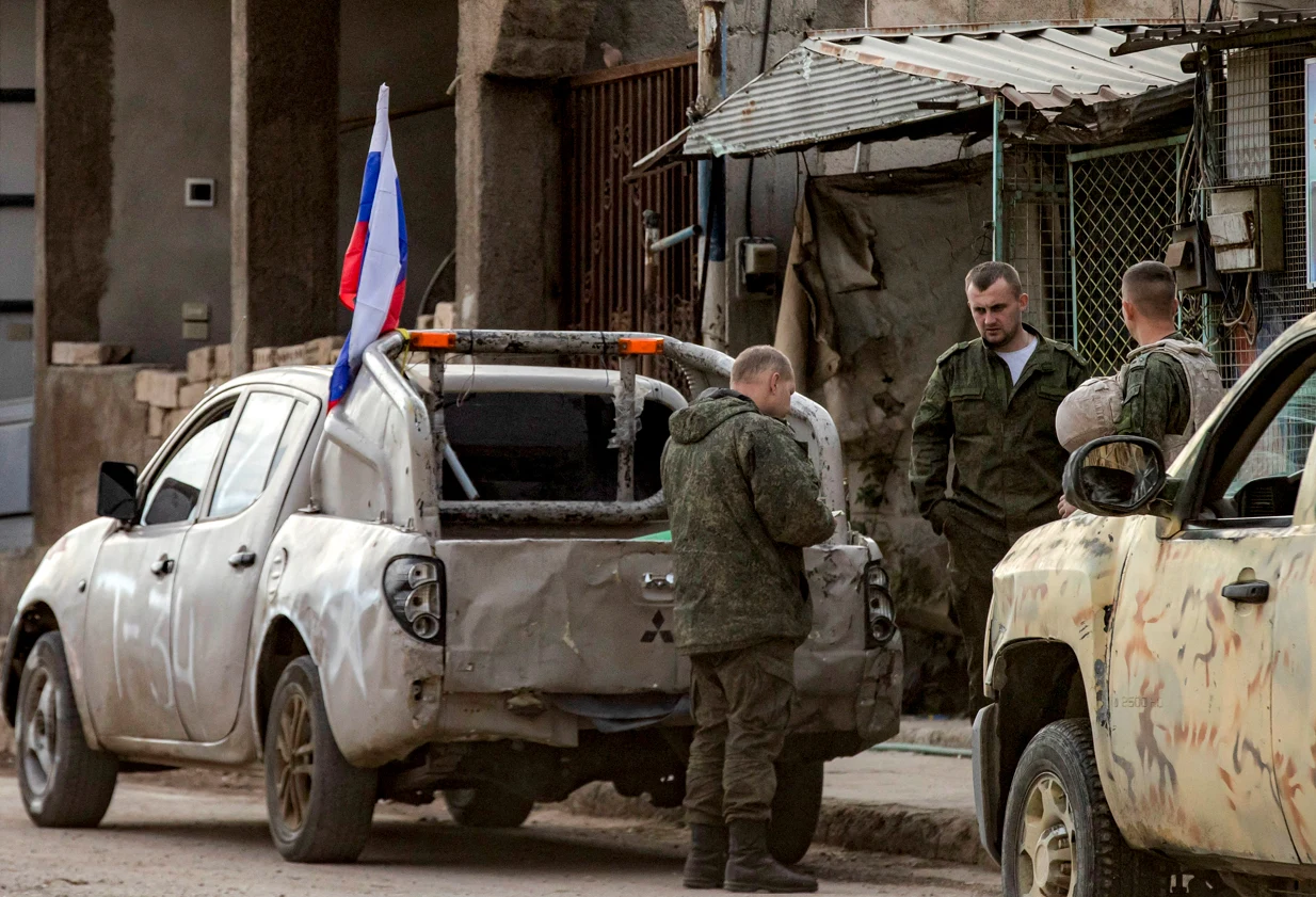 Czterech żołnierzy w mundurach stoi obok dwóch zniszczonych pojazdów, z których na jednym znajduje się flaga Rosji, otoczenie stanowią zrujnowane zabudowania i prowizoryczny punkt kontrolny.