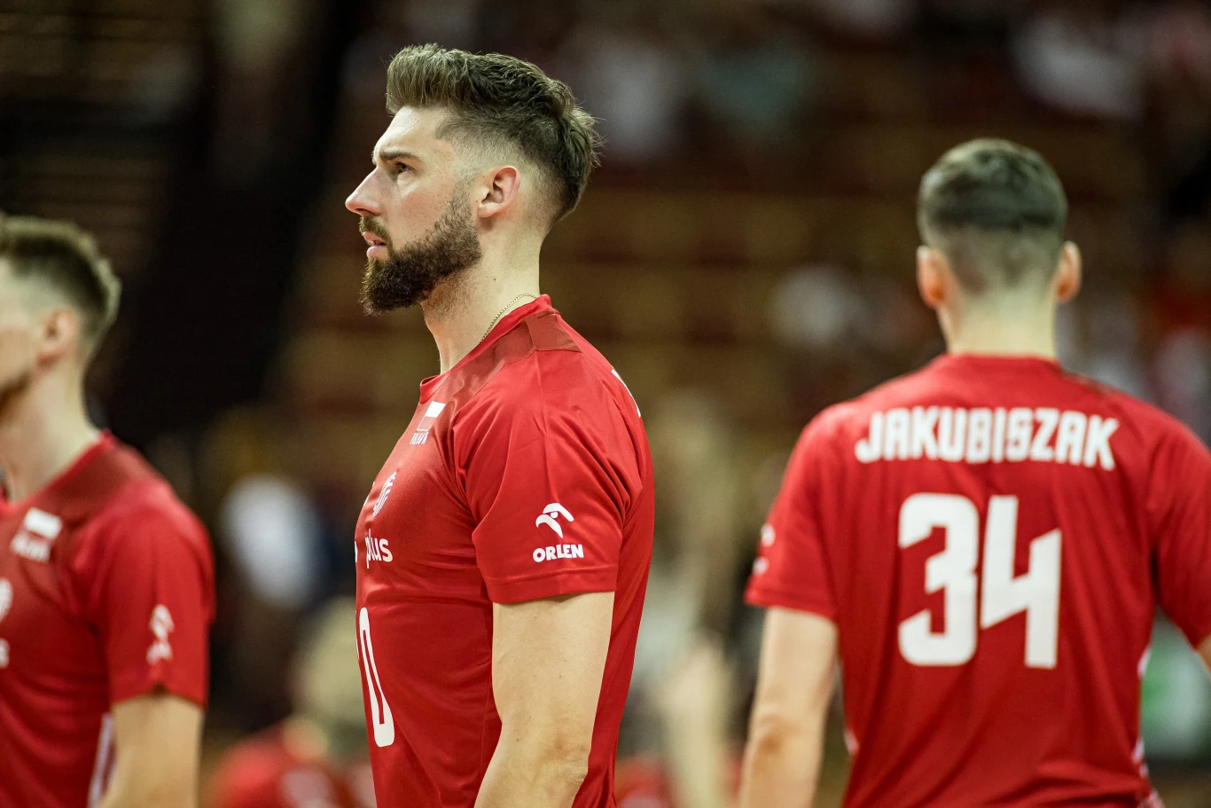 Dwóch siatkarzy w czerwonych koszulkach reprezentacji Polski stoi pośród rozgrywanego meczu, jeden widoczny z profilu z numerem 10, drugi od tyłu z numerem 34, sylwetki na tle rozmytej widowni.