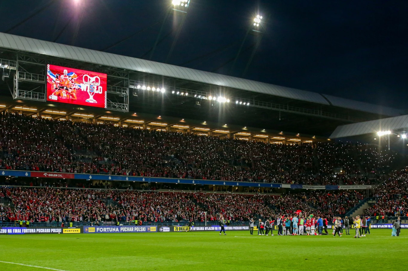 Tłum kibiców na wypełnionym stadionie podczas meczu piłkarskiego, oświetlone trybuny, piłkarze i obsługa techniczna na murawie, na ekranie widoczna grafika związana z pucharem piłkarskim.