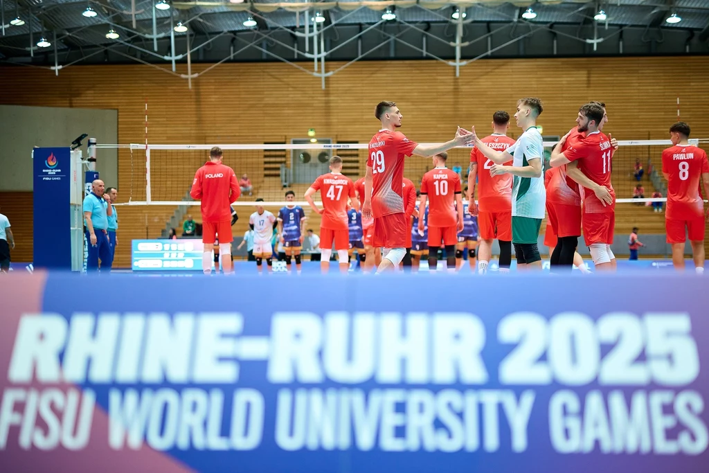 Uniwersjada 2025 Siatkarska drużyna w czerwonych strojach na hali sportowej świętuje udaną akcję, zawodnicy wymieniają gesty gratulacji, w tle drugi zespół ubrany na niebiesko oraz siatka. W pierwszym planie widoczna tablica promująca FISU World University Games Rhine-...