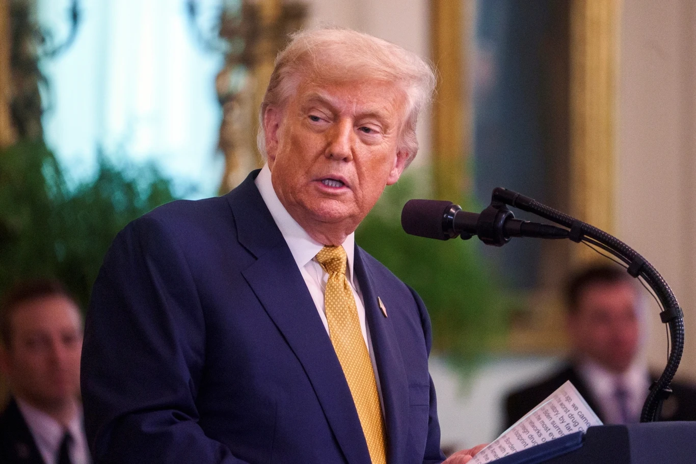 Prezydent USA Donald Trump Mężczyzna w granatowym garniturze i żółtym krawacie stoi przy mównicy z mikrofonem, trzymając notatki; w tle widoczne dekoracje i inne osoby w rozmyciu.