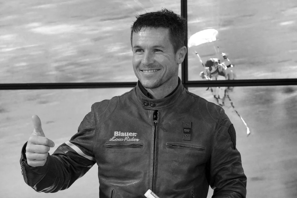Felix Baumgartner Uśmiechnięty mężczyzna w skórzanej kurtce, stojący na tle dużego okna, za którym widać spadochroniarza lądującego na polu.