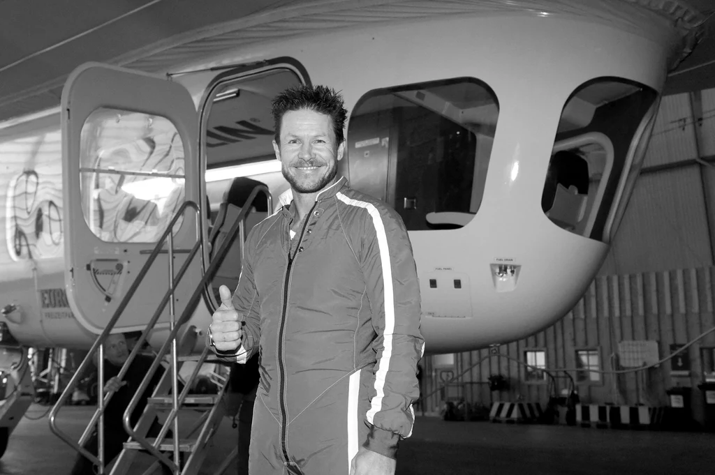 Felix Baumgartner Mężczyzna w kombinezonie lotniczym pozuje z uniesionym kciukiem na tle niewielkiego samolotu pasażerskiego na płycie lotniska.