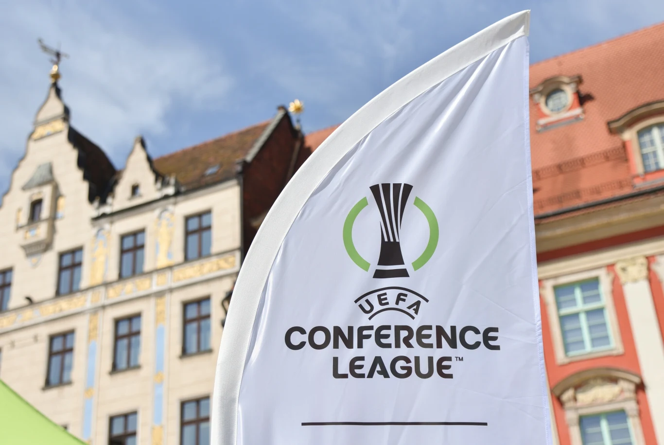 HJK pozostaje w grze o ścieżkę ligową Ligi Konferencji Flaga UEFA Conference League na pierwszym planie, w tle zabytkowe fasady kamienic z czerwonymi dachami i niebieskim niebem.