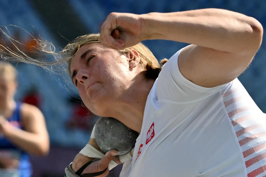Zuzanna Maślana Zbliżenie na zawodniczkę w trakcie pchnięcia kulą, skupienie widoczne na jej twarzy, sportowa koszulka z polskim godłem, w tle rozmyte trybuny stadionu.