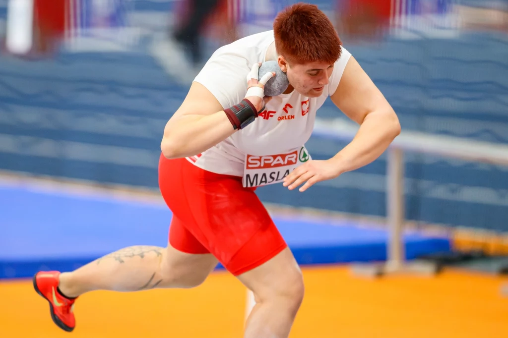 Zuzanna Maślana Lekkoatleta w polskim stroju sportowym w trakcie pchnięcia kulą, ujęcie dynamiczne podczas zawodów lekkoatletycznych, wyraźnie widoczna koncentracja i siła sportowca.
