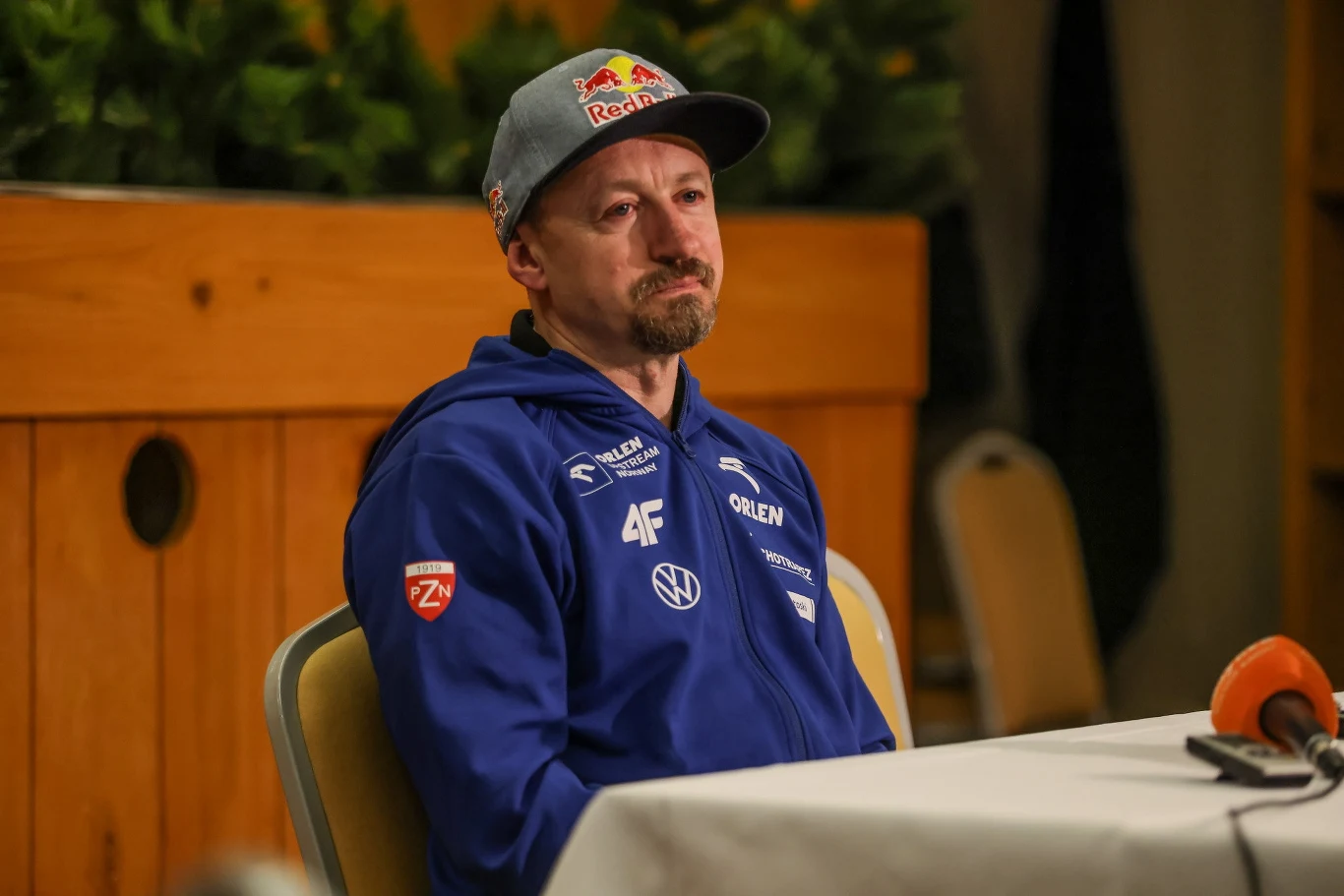 Mężczyzna w sportowej bluzie i czapce z daszkiem siedzący przy stole konferencyjnym, z poważnym wyrazem twarzy, w tle drewniana ścianka i rośliny ozdobne.