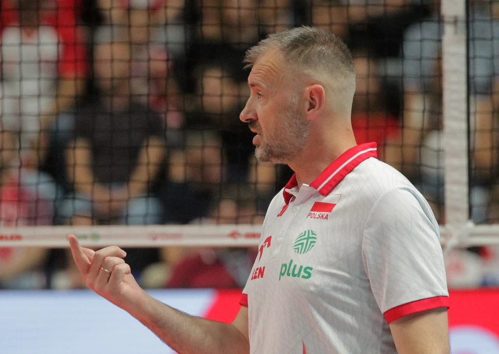 Nikola Grbić Trener siatkarski w białej koszulce z czerwonym kołnierzykiem, stojący przy siatce na tle rozmazanej publiczności, wykonujący gest ręką w kierunku boiska.