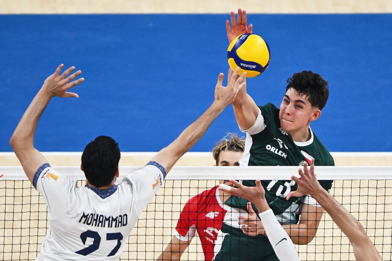 Dynamika meczu siatkówki, dwóch zawodników rywalizuje przy siatce o zdobycie punktu, piłka znajduje się tuż nad dłońmi graczy, w tle widać fragment boiska i kolejnego siatkarza.