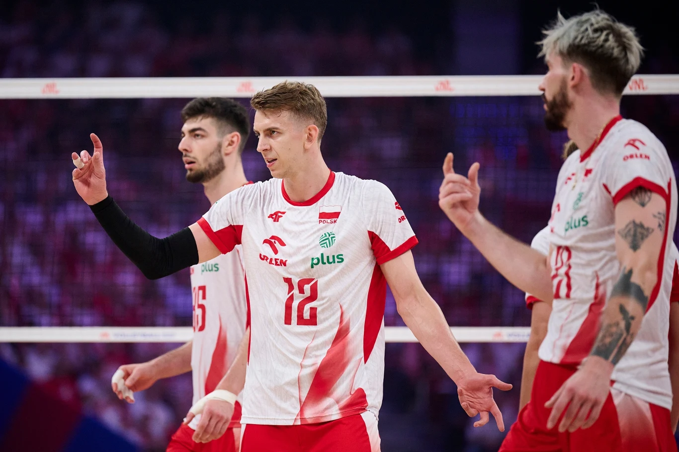 Trzech siatkarzy polskiej reprezentacji w biało-czerwonych strojach podczas meczu, jeden z nich pokazuje gest ręką, wszyscy skupieni na rozgrywce, w tle siatka i zamazane trybuny.