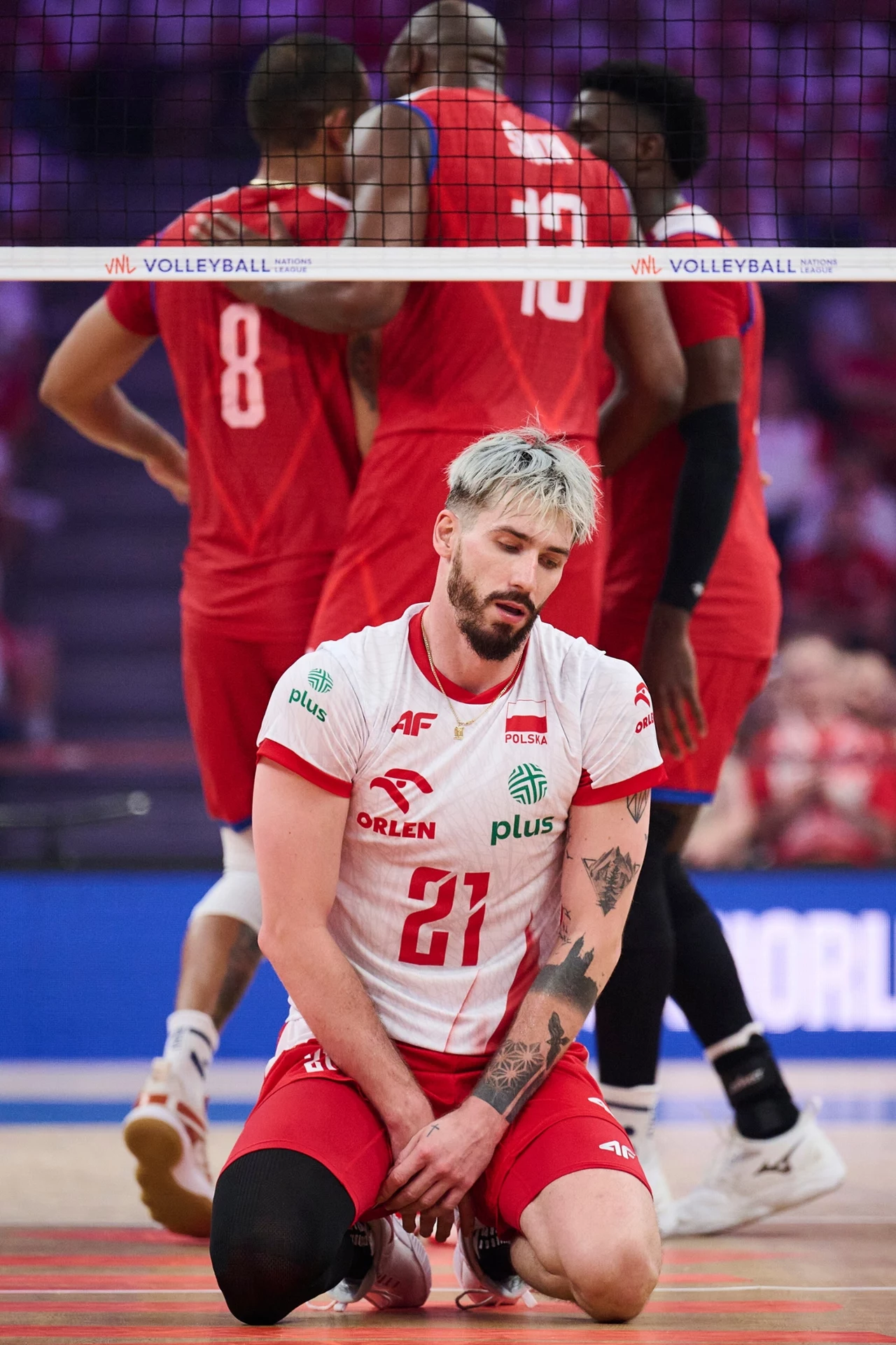 Tomasz Fornal Siatkarz w biało-czerwonym stroju z numerem 21 klęczy na parkiecie, w tle trzech zawodników drużyny przeciwnej ubranych w czerwone stroje, za siatką, w hali wypełnionej kibicami.