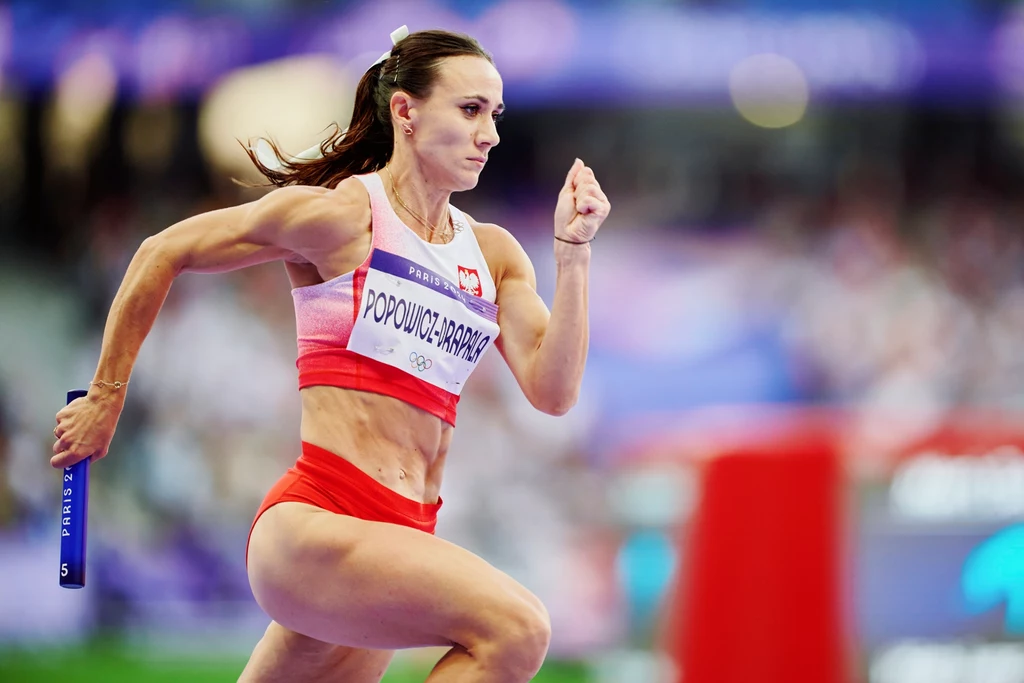 Marika Popowicz-Drapała Zawodniczka biegnąca z pałeczką sztafetową na tle stadionu, ubrana w sportowy strój biało-czerwony, widoczna energia i skupienie na twarzy.