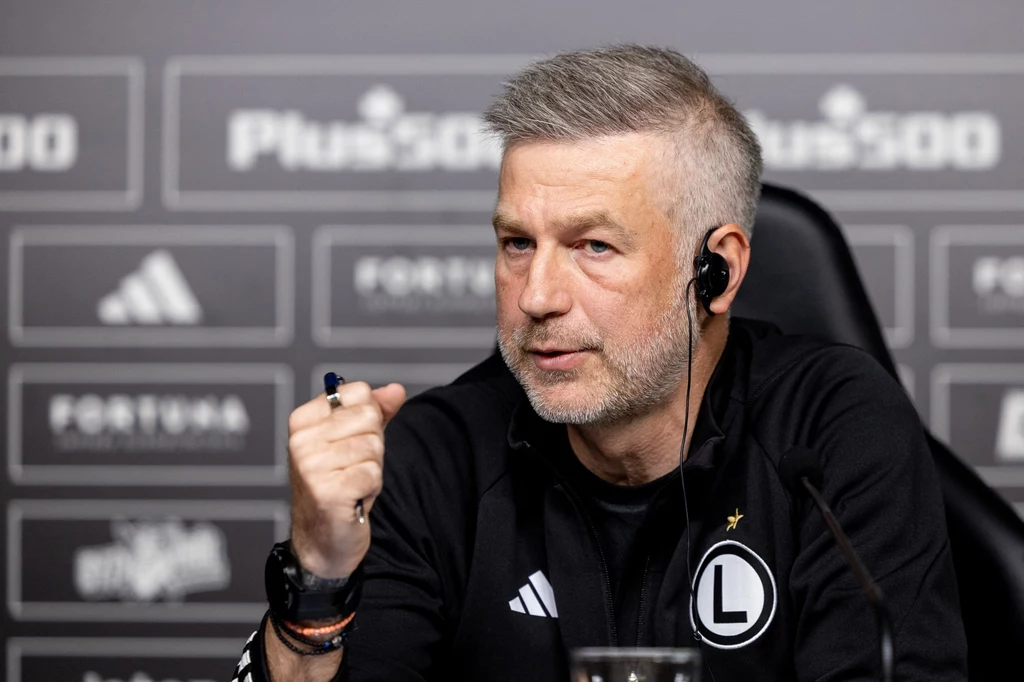 Edward Iordanescu - trener Legii Warszawa Mężczyzna w średnim wieku z krótko ostrzyżonymi, siwymi włosami siedzi przy stole konferencyjnym, ubrany w sportową bluzę z logiem klubu Legia Warszawa, z mikrofonem i słuchawką w uchu, zaciska dłoń w geście podkreślenia wypowiedzi.