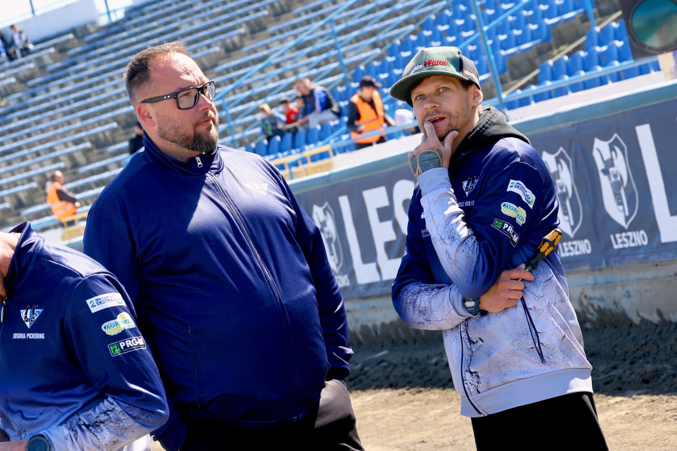 Sławomir Kryjom w rozmowie z Januszem Kołodziejem Dwóch mężczyzn w sportowych kurtkach stoi na tle pustych trybun stadionu. Jeden z nich trzyma w ręku marker i ma zadumaną minę, podczas gdy drugi patrzy w stronę kamery w okularach. W oddali widoczni są inni pracownicy stadionu oraz niebieskie fotele d...