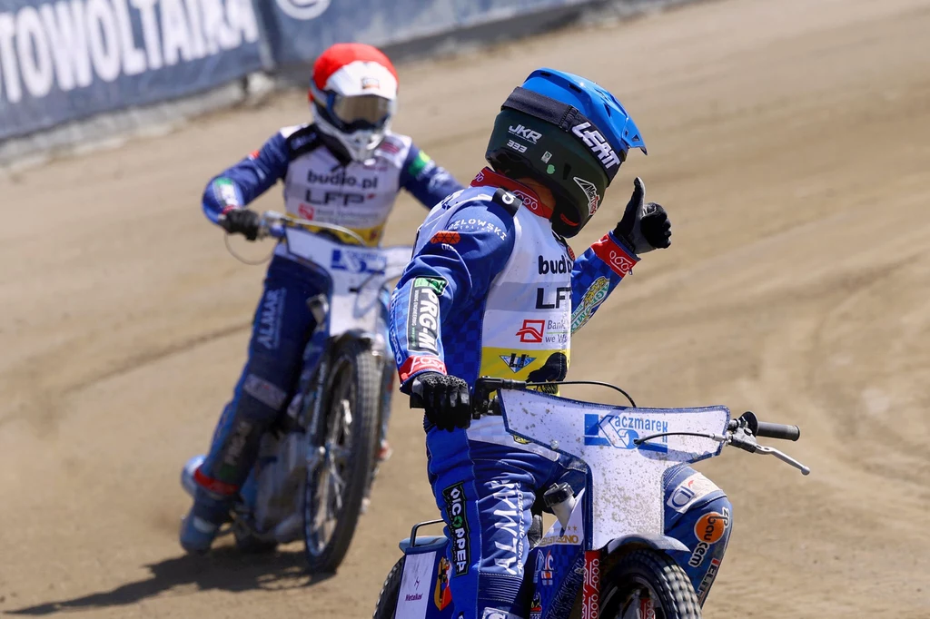 Janusz Kołodziej i Nazar Parnicki Dwóch żużlowców w kolorowych kombinezonach i kaskach jadących na motocyklach na torze, jeden z nich pokazuje gest kciuka uniesionego do góry.