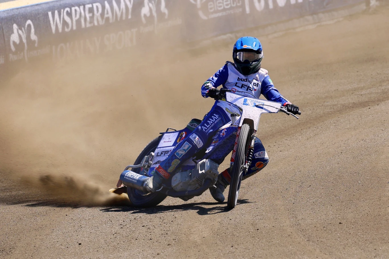Zawodnik w niebieskim kombinezonie i kasku wykonuje dynamiczny zakręt na motocyklu żużlowym, wzbijając za sobą chmurę kurzu na torze, wokół widoczne reklamy oraz pobocze toru.