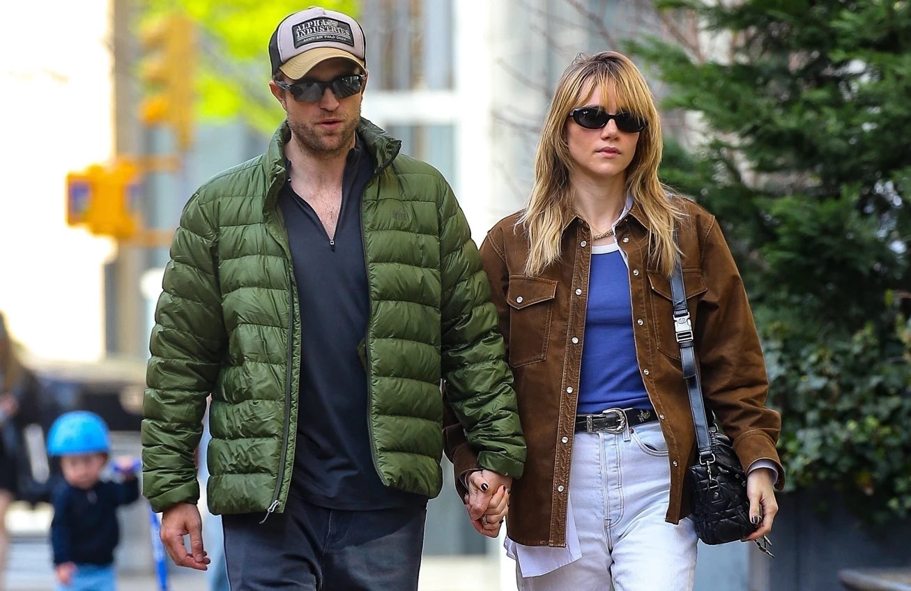 Robert Pattinson i Suki Waterhouse Para spaceruje trzymając się za ręce, oboje noszą okulary przeciwsłoneczne i casualowe ubrania, w tle widać nieostry fragment miasta oraz przechodzące dziecko na hulajnodze w kasku.