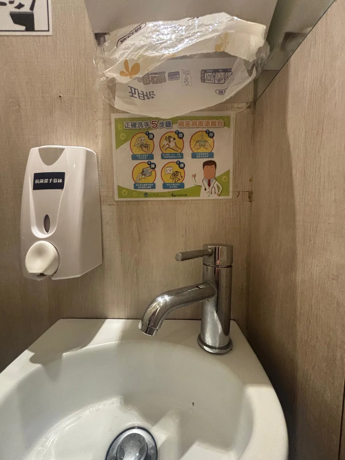Umýwalka z chromowaną baterią, dozownik mydła zamontowany na ścianie po lewej stronie, powyżej umywalki instrukcja mycia rąk w języku azjatyckim z ilustracjami, na górze widoczna plastikowa osłona na deski sedesowe.