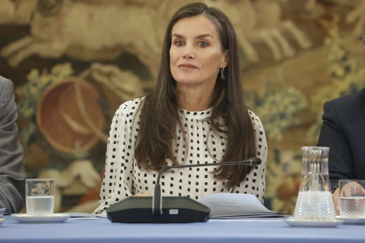 Królowa Letizia zachwyciła w sukience, którą ma w swojej szafie już od 7 lat Kobieta o długich, brązowych włosach w białej bluzce w czarne kropki siedzi przy stole konferencyjnym, przed nią znajduje się mikrofon, szklanka z wodą i karafka, w tle dekoracyjna tapiseria.
