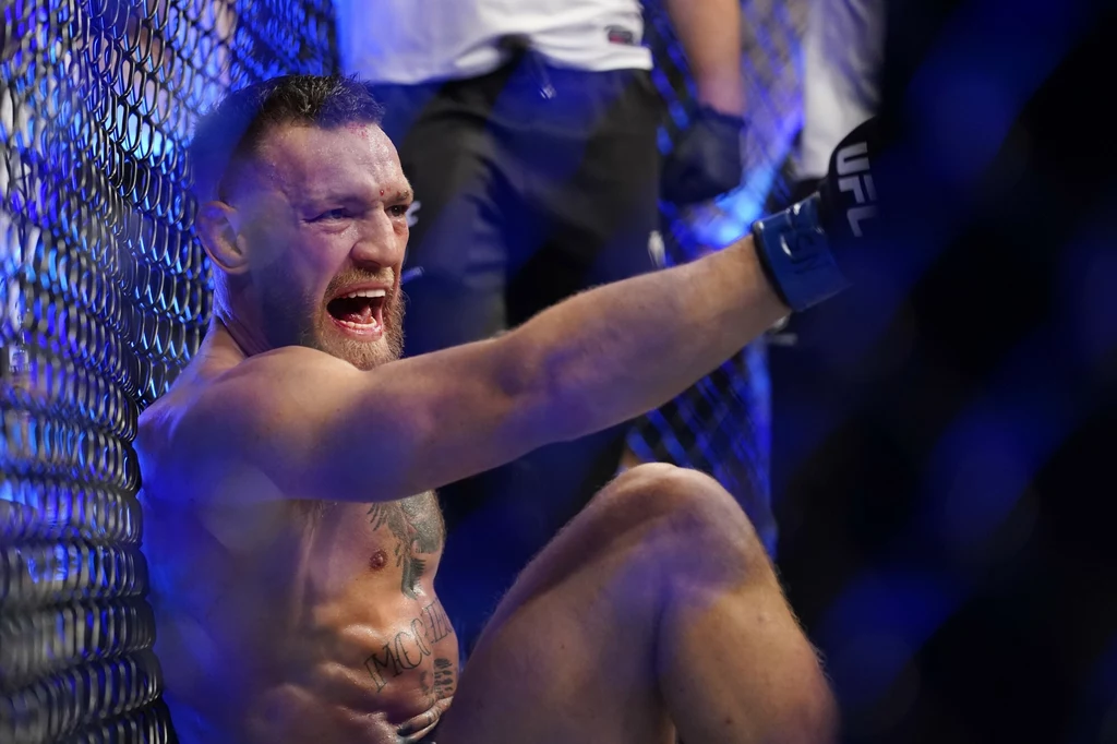 Conor McGregor zakończył ostatnią walkę w UFC z kontuzją Zawodnik MMA siedzi przy siatce oktagonu z wyrazem bólu lub frustracji na twarzy, w tle widoczny fragment areny i sylwetki osób z obsługi walki.